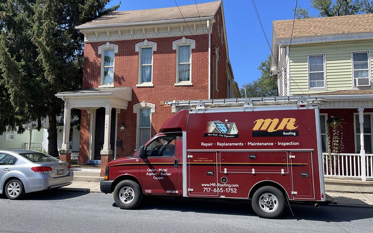 A red MR Roofing service van parked in front of historic brick and clapboard houses.