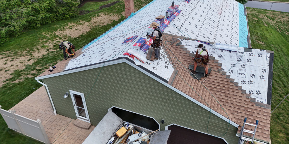 Aerial view of roofers installing shingles over GAF underlayment on a green sided house.