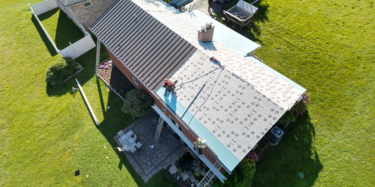 Aerial view of workers installing GAF underlayment on a large roof partly finished with metal panels.